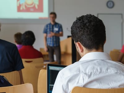 Ein Mann sitzt mit dem Rücken zum Betrachter, im Hintergrund hält ein anderer Mann eine Präsentation